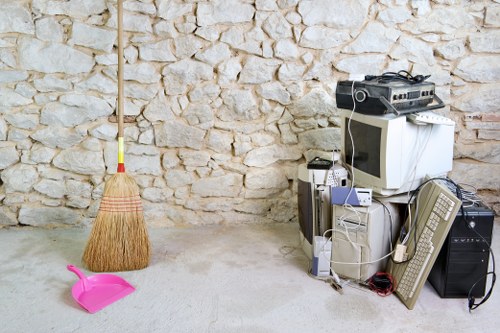 A collection of discarded household electronic devices and appliances, including a CRT computer monitor, a desktop tower, an old-style keyboard, a box-shaped printer or scanner, and various cables, all stacked on the floor in front of a light-colored, rough-textured stone wall. To the left, there is a wooden-handled broom with a yellow grip leaning against the wall, and a small pink dustpan with a handle resting on the ground nearby. The setting appears to be a garage or storage area, with a neutral, unpainted concrete floor. This scene, associated with waste disposal services in Fulham, reflects the process of clearing out electronic waste for removal or recycling, a service offered by Commercial Waste Fulham in the local area.