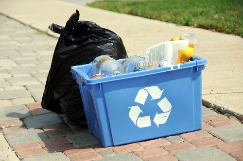 A blue plastic recycling bin placed on a paved sidewalk near a grassy area, filled with clear plastic bottles, a cardboard container, and other waste items. Next to it is a large black rubbish bag that is tied at the top, containing disposed materials. The background shows a curb and street surface, indicating an outdoor setting typical for waste collection or rubbish removal in Fulham. The scene reflects typical rubbish and recycling collection in an urban environment, with the bin and bag positioned for pickup by waste management services such as Commercial Waste Fulham, which operates in the SW6 postcode district of London.