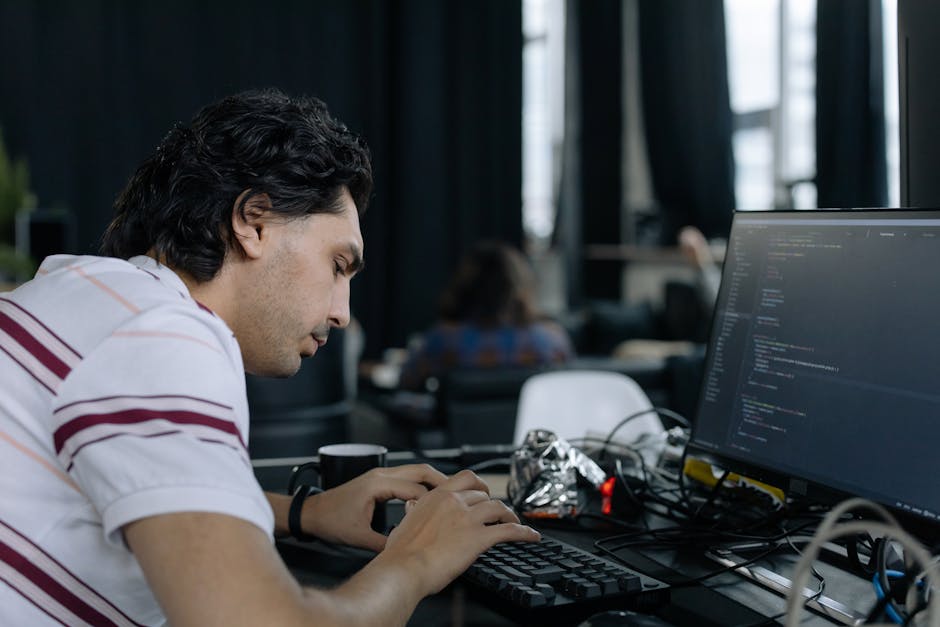 A person's hand, wearing a wristwatch with a black strap and a round dial, is positioned over a silver laptop keyboard, which displays lines of code on a dark background with color-coded syntax highli